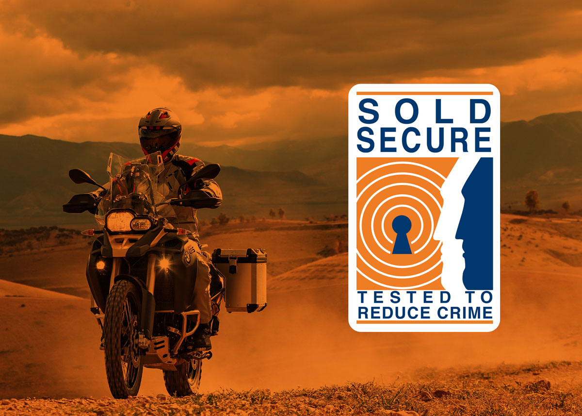 Person riding a motorcycle on a desert road with a 'Sold Secure' logo overlay.